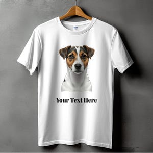 May include: White t-shirt featuring a Jack Russell Terrier portrait with brown, black, and white markings. The text "Your Text Here" is printed below the dog image. The shirt is hanging on a wooden hanger.