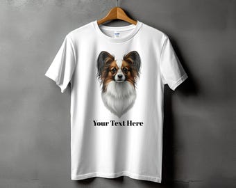 Puede incluir: Camiseta blanca con una ilustración detallada de la cara de un perro Papillon. El perro tiene pelo marrón, negro y blanco. El texto "Your Text Here" está impreso debajo de la imagen del perro. La camiseta está colgada en una percha de madera.