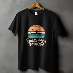 以下が含まれることがあります： カヌーを漕ぐ人のシルエットとレトロな夕焼けのグラフィックがプリントされた黒いTシャツ。グラフィックの下に「Paddle More, Worry Less」というテキストがプリントされています。
