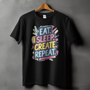 May include: Black t-shirt with a colorful graphic design that says "Eat. Sleep. Create. Repeat." The graphic features colorful pencils and paintbrushes.