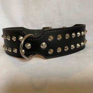 Puede incluir: Collar de perro de cuero negro con tachuelas de pirámide plateadas. El collar tiene un anillo en D y una hebilla.