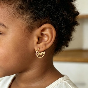 May include: Gold-colored hoop earring with the name "Chase" in a cursive font. The earring is worn on the ear of a person with dark skin and curly hair. The earring is a simple, elegant design.