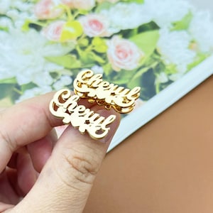May include: Two gold-colored name pins, each spelling "Cheryl" in a cursive font. The pins are displayed against a backdrop of flowers. The pins are likely jewelry or accessories.