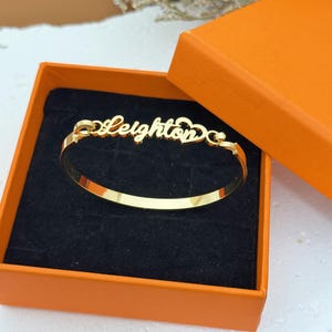 May include: A gold-colored bangle bracelet with the name "Leighton" in a cursive font, presented in an orange gift box with a black interior. The bracelet has a heart-shaped design element.