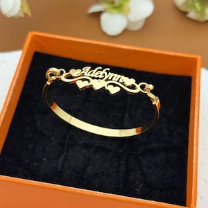 May include: A gold-coloured bangle bracelet inscribed with the name "Adelynn" in a cursive font. The bracelet has heart-shaped details and is displayed in an orange box with a black interior. The bracelet is a piece of jewellery.