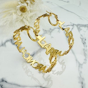 May include: A pair of gold hoop earrings with the word "LAROMA" spelled out in large letters. The earrings are made of metal and have a simple, modern design.