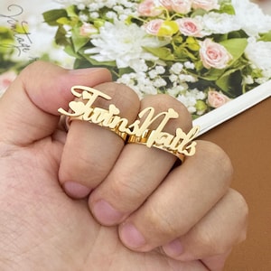 Puede incluir: Anillo doble para dedos de color dorado con el nombre "Twins Nails" en fuente cursiva. El anillo presenta pequeños detalles de corazón y se usa en dos dedos. El fondo incluye flores.