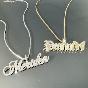 Puede incluir: Dos collares de plata y oro con los nombres personalizados "Meriden" y "Peanut" con un dije de mariposa. Los nombres están engastados con diamantes de imitación transparentes.