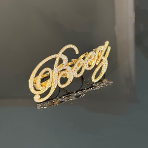 Puede incluir: Un anillo dorado para varios dedos que muestra la palabra "Boog" en una fuente cursiva. La superficie de las letras está incrustada con pequeñas piedras claras y brillantes que captan la luz, creando un aspecto luminoso y reflectante.