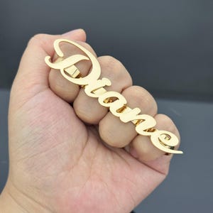 May include: A gold-colored ring spelling out the name "Diane" in a cursive font. The ring is held in a hand against a neutral background. The ring is a personalized jewelry item.