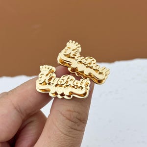 May include: Two gold-colored, layered nameplate pins. Each pin features the name "Fly Boy" in a cursive font, topped with a small crown. The pins are displayed against a brown background.