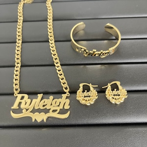 May include: Gold necklace, bracelet, and earrings with the name "Ryleigh" spelled out on each piece. The necklace has a curb chain and a large nameplate pendant with a heart design. The bracelet is a simple bangle with the name on it. The earrings are hoop earrings with the name on them.