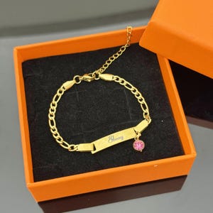 Peut inclure: Un bracelet doré avec une plaque nominative gravée du nom "Ebony". Le bracelet présente un motif de chaîne et un petit charme en forme de pierre précieuse rose. Présenté dans un coffret cadeau orange et noir.