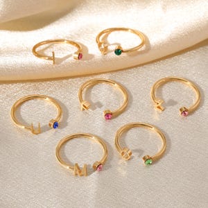 Peut inclure: Anneaux dorés avec des initiales et des pierres précieuses colorées. Les bagues présentent des lettres comme "T", "U", "M" et "O", chacune associée à une gemme de couleur différente, notamment rose, vert et bleu. Les anneaux sont présentés sur une surface crème douce.
