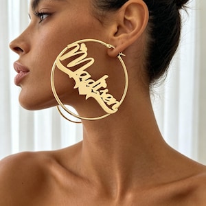 May include: Gold-coloured hoop earrings featuring the name "Madison" in a cursive font. The earrings are large and circular, with a heart-shaped detail. The earrings are worn on a person's ear.