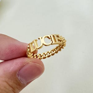 Puede incluir: Anillo de oro con el nombre "LUCIE" escrito en letras mayúsculas en una banda de cadena.