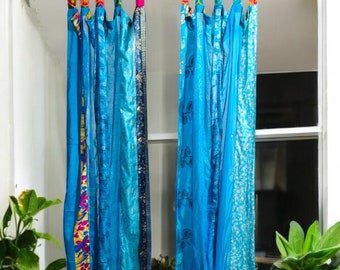 May include: Two blue sari curtains hang in a window, featuring diverse blue patterns and textures. The curtains are suspended from a metal rod. The top of the curtains have colourful patterned fabric. Potted plants sit on either side of the window.