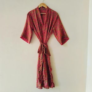 May include: A pink floral patterned robe hanging on a wooden hanger. The robe has a V-neck and a tie at the waist. The floral pattern includes shades of green and gold. The robe is made of a lightweight fabric.