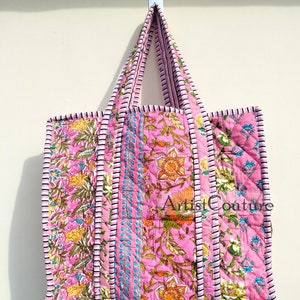 Puede incluir: Un bolso tote acolchado rosa con estampados florales en amarillo, naranja y verde. El bolso tiene un asa y ribete a rayas. El texto "Artist Couture" es visible en el bolso.