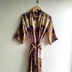 May include: A floral patterned robe in shades of beige, purple, and gold hangs on a wooden hanger. The robe features a tie closure at the waist and a repeating floral design. The sleeves are three-quarter length, and the robe is made of a lightweight fabric.