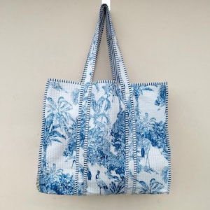 May include: A white tote bag with a blue tropical print of palm trees and birds. The bag has blue striped trim and handles. It is hanging against a neutral wall.
