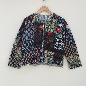 May include: A patchwork jacket with a round neckline and long sleeves. The jacket features a variety of floral and geometric patterns in black, white, red, orange, and green. The jacket is hanging on a wooden hanger against a neutral background.