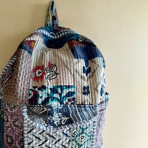 Könnte beinhalten: Ein Patchwork-Stepprucksack mit verschiedenen blauen, grauen und weißen Blumenmustern. Der Rucksack hat eine einzelne Tasche auf der Vorderseite und zwei Träger.
