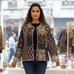 May include: A black quilted jacket with a floral patchwork design. The jacket has a round neckline and is trimmed with a white and black striped fabric. The person is wearing blue jeans and tan ankle boots.