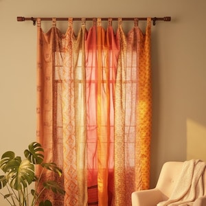 Puede incluir: Cortinas transparentes de color naranja y amarillo con diseños, colgadas de una barra de madera. Las cortinas presentan varios patrones y texturas. Una planta en maceta y un sillón beige con una manta están en primer plano.