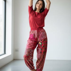 以下が含まれることがあります： 赤い半袖トップスとパッチワークパンツを着用した人物。パンツは赤、ピンク、バーガンディの色合いで様々な模様が施されています。人物の両腕は頭上に上げられています。