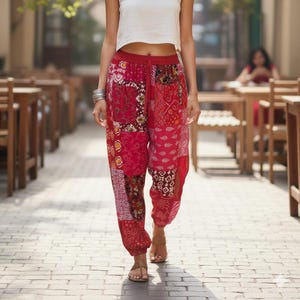 May include: A woman in a white crop top and red patchwork trousers. The trousers display various patterns and shades of red. She is walking on a brick-paved walkway, with tables and chairs in the background.