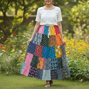 Puede incluir: Una mujer con una blusa blanca y una falda larga de patchwork. La falda presenta una variedad de cuadrados de tela coloridos en tonos de rosa, azul, verde, amarillo y naranja. La falda llega hasta los tobillos.