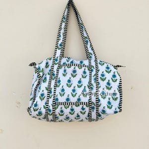 Könnte beinhalten: Weiße Baumwoll-Tragetasche mit blauem und grünem Blumendruck und schwarz-weiß gestreiftem Besatz. Die Tasche hat zwei Henkel und der Text "Artist Couture" ist auf der Vorderseite sichtbar.