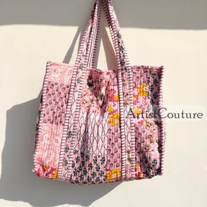 May include: Pink quilted tote bag with floral and geometric patterns. The bag has sturdy handles and is hanging against a light-colored wall. The text "Artist Couture" is visible on the bag.