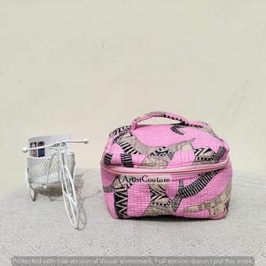 May include: A pink cosmetic bag with a handle and a zipper, featuring a pattern of black and tan shapes. The bag has the text "Artist Couture" on the front. A white metal bicycle-shaped decorative item is to the left.