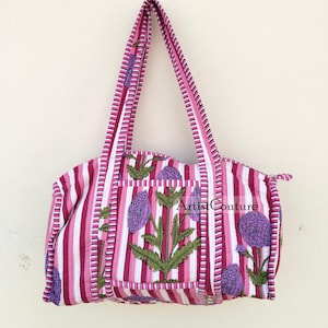 Puede incluir: Bolso de mano de rayas rosa y blancas con estampado floral de flores moradas y hojas verdes. El bolso tiene una sola correa y un pequeño bolsillo en la parte delantera. El texto "Artist Couture" es visible en el bolso.