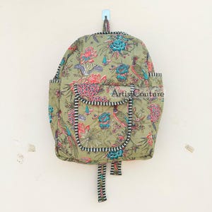 Puede incluir: Una mochila acolchada verde con estampado floral con pájaros y flores. La mochila tiene cierre de cremallera y un pequeño bolsillo en la parte delantera. La mochila tiene un ribete negro y blanco alrededor de los bordes.