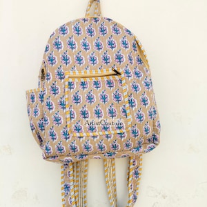 May include: A beige backpack featuring a repeating floral design in pink and teal. It has a front zipped pocket, adjustable straps, and the text "Artist Couture". The bag is suspended on a wall.