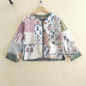 Puede incluir: Una chaqueta acolchada blanca con un diseño de patchwork que presenta patrones florales y geométricos en tonos de rosa, azul, verde y amarillo. La chaqueta tiene un escote en V, mangas largas y cierre de botones. La tela tiene un ribete a rayas en los bordes.
