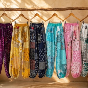 Peut inclure: Six pantalons amples avec taille et chevilles élastiquées, chacun présentant un motif patchwork distinct en violet, jaune, bleu marine, turquoise, rose et blanc, suspendus à des cintres en bois sur une branche horizontale.