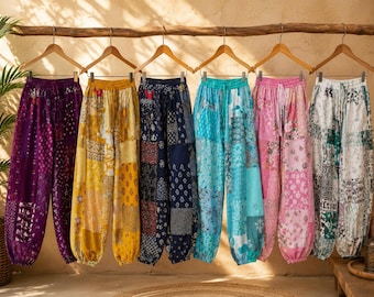 Puede incluir: Seis pantalones holgados con cinturillas y puños elásticos, cada uno con un patrón de patchwork distinto en morado, amarillo, azul marino, turquesa, rosa y blanco, colgados de perchas de madera en una rama horizontal.