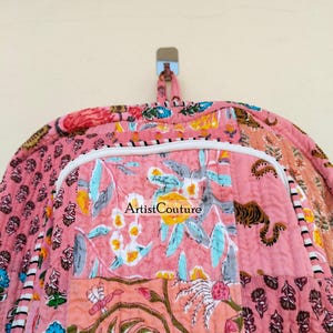 May include: A pink quilted backpack with a floral patchwork design. The backpack features a white zipper and a handle at the top. The design includes a tiger print and the text "Artist Couture".