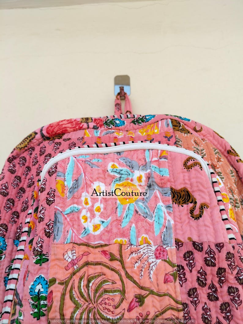 May include: A pink quilted backpack with a floral patchwork design. The backpack features a white zipper and a handle at the top. The design includes a tiger print and the text "Artist Couture".