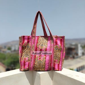 May include: A pink tote bag featuring a tiger stripe design and vertical pink stripes. The bag has red and white striped handles and the text "Artist Couture". The bag is made of a quilted fabric.
