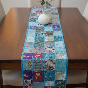 Könnte beinhalten: Ein Patchwork-Tischläufer mit blauem Rand und einer Vielzahl farbenfroher Stoffquadrate. Eine weiße Vase mit grünen Zweigen steht auf dem Läufer, der auf einem dunklen Holztisch liegt. Das Design des Läufers umfasst Blumen- und Paisley-Muster.