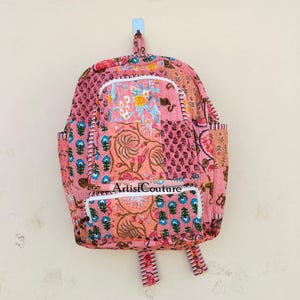 May include: Pink patchwork backpack with floral and paisley patterns. Features a front pocket and side pockets. The text "Artist Couture" is visible on the front pocket. The backpack is hanging on a wall.
