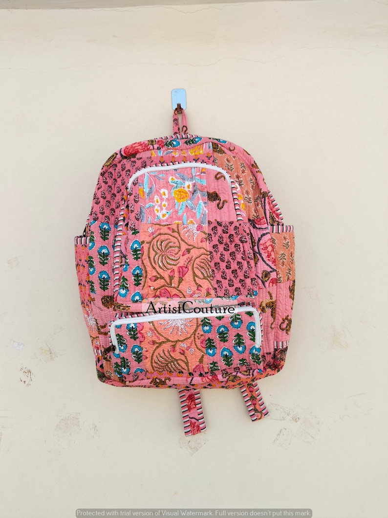 May include: Pink patchwork backpack with floral and paisley patterns. Features a front pocket and side pockets. The text "Artist Couture" is visible on the front pocket. The backpack is hanging on a wall.