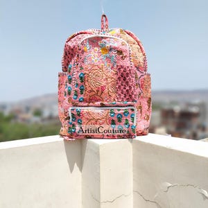 Op de afbeelding: Roze gewatteerde rugzak met bloemen- en paisley-patronen, met een voorvak met de tekst "Artist Couture". De rugzak heeft een handvat aan de bovenkant en zijvakken. Het ontwerp bevat blauwe en witte accenten.