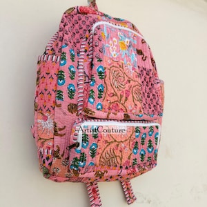 May include: Pink quilted backpack with floral and paisley patterns. Features two front pockets and adjustable straps. The text "Artist Couture" is visible on the front pocket. The backpack is hanging on a wall.