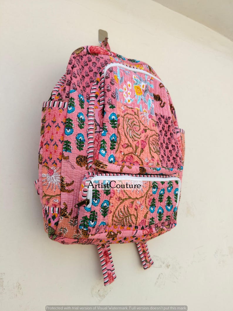 May include: Pink quilted backpack with floral and paisley patterns. Features two front pockets and adjustable straps. The text "Artist Couture" is visible on the front pocket. The backpack is hanging on a wall.